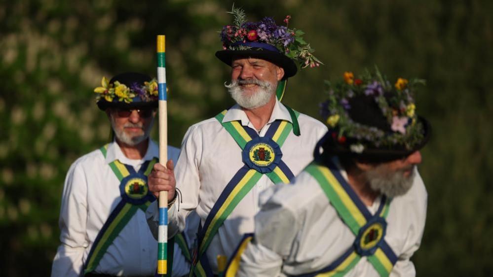 Three men wearing Morris dancer outfits. One is carrying a pole decorated with colourful paint. They are all wearing cross sashes of green, yellow and blue and hats covered with flowers and leaves.