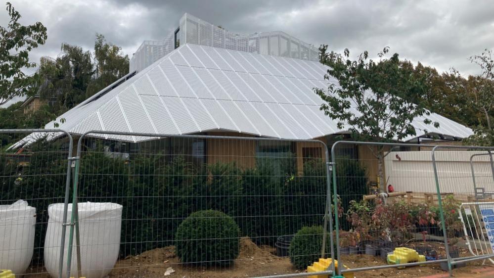 A building behind fencing. The roof is grey and shaped like a hut. There are trees in front.