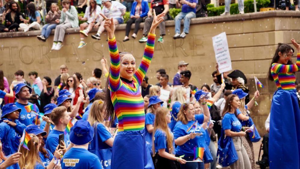 A Pride parade in a city. A woman is dressed in blue trousers and a striped multi-colour top. She has both her arms in the air and is smiling a towards the camera. A group of people walk behind her in the parade all dressed in blue tops and holding Pride flags.