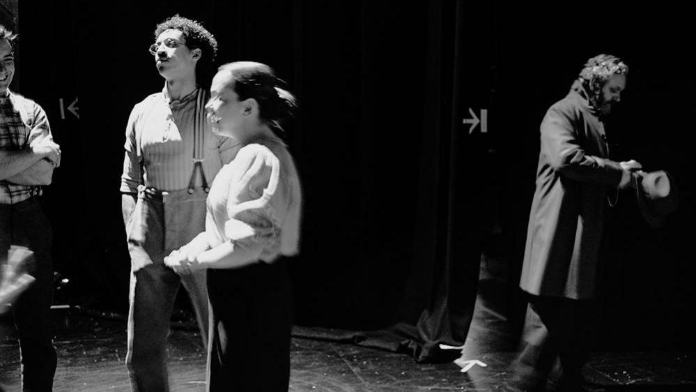 Michael Sheen, wearing a long coat, is seen to the right of the screen as a group of three people are seen talking to the left. There are arrows on the floor and an arrow sign on a door which suggest they are on a stage.