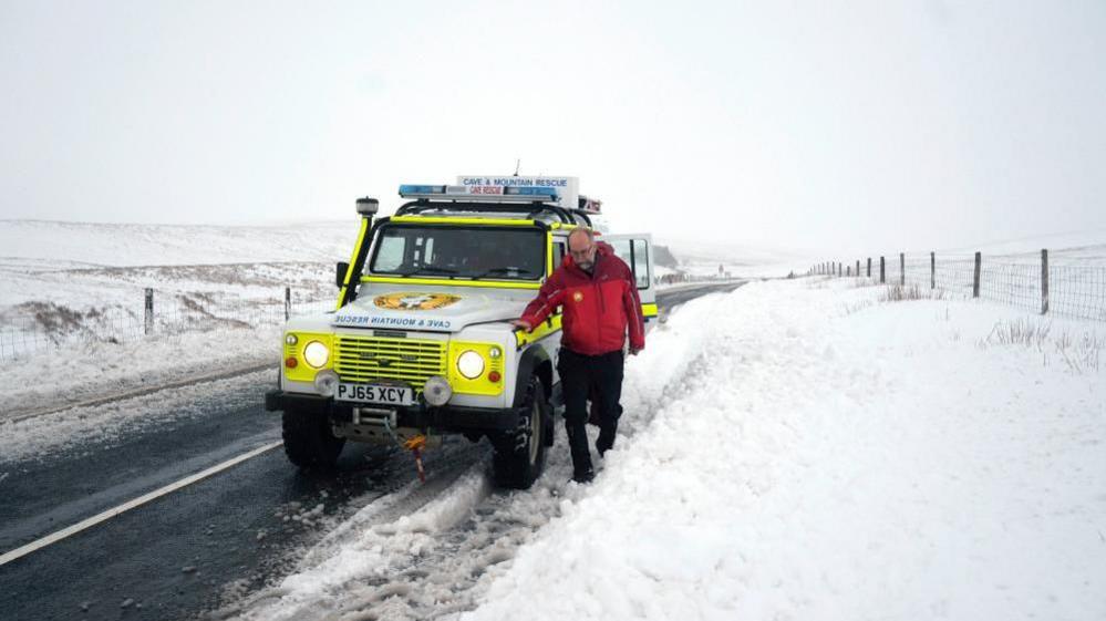 A snowy road on the moors with a mountain rescue van and worker walking down the road 