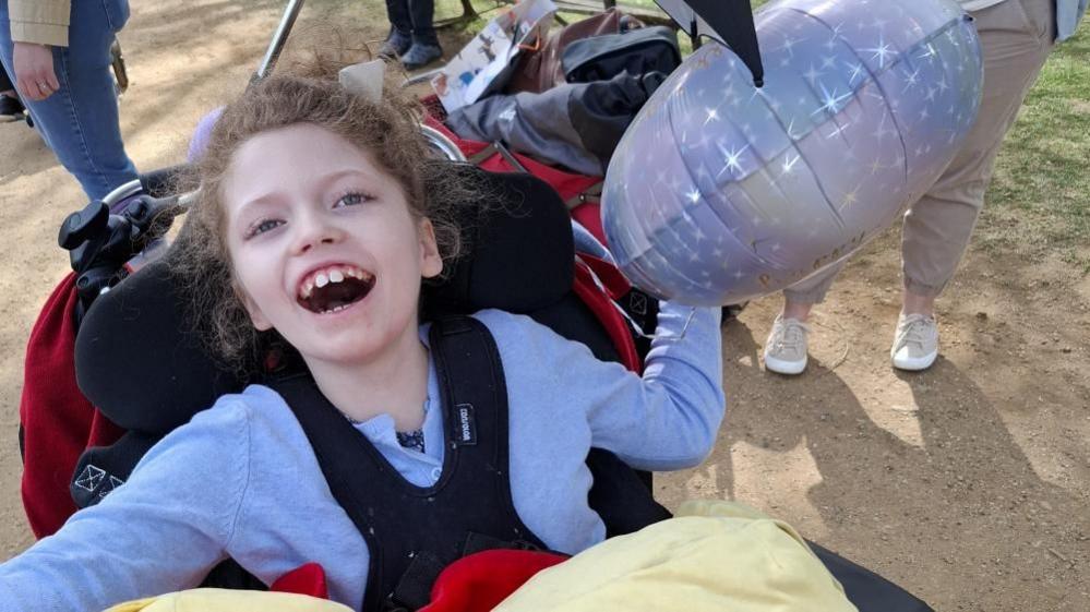Addy Clarke from Swindon, Wiltshire. She is smiling whilst in a specialist wheelchair and holding a balloon.