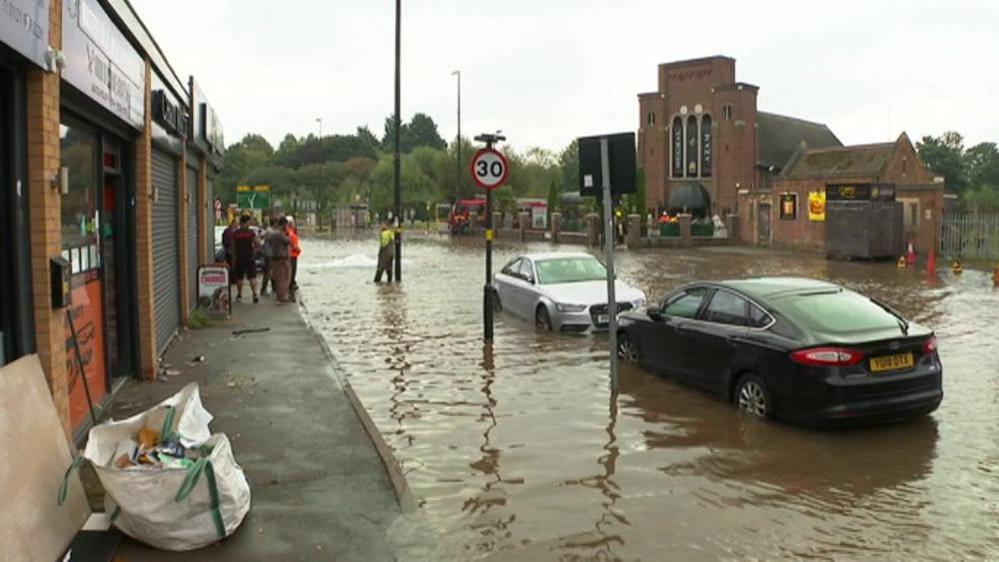 Stratford Road, Birmingham, with two abandoned cars under inches of water. A group of residents looks on from a pavement with a man in wellies in the road. A restaurant is in the background.