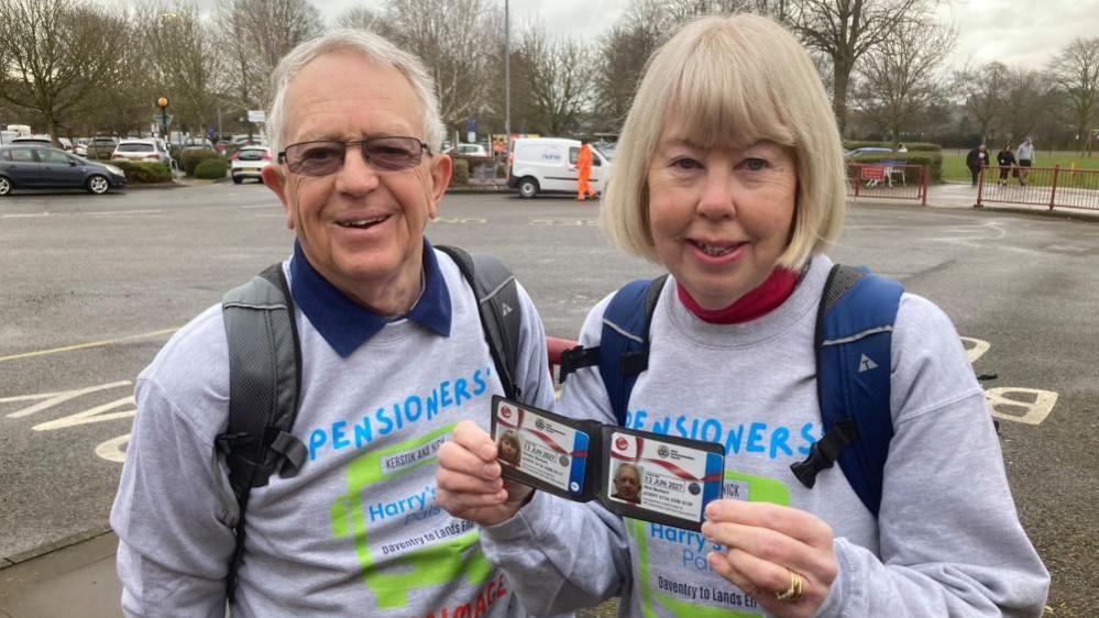 Nick and Kerstin wearing grey sweatshirts labelled "Pensioners' Pilgrimage" and "Harry's Pals". Nick has short white hair while Kerstin has short blonde hair. They are both wearing backpacks. Kerstin is showing their bus passes to the camera. There is a large bus turnaround area in the background.