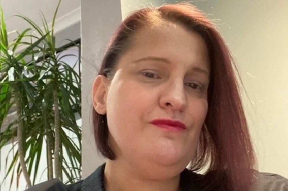 Jo smiles at the camera. She has a red bob hair cut and wears a black top. A plant is in the background.