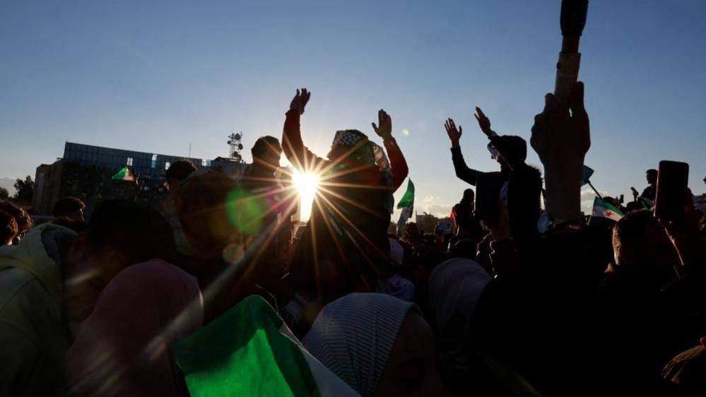 The sun is setting and you can see people in a big crowd holding onto Syrian flags as they cheer.