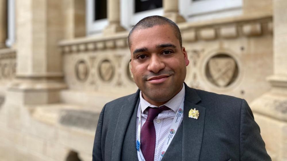 James Hill with short dark hair smiling at the camera outside a stone building wearing a dark suit and red tie with a council crest on the right lapel