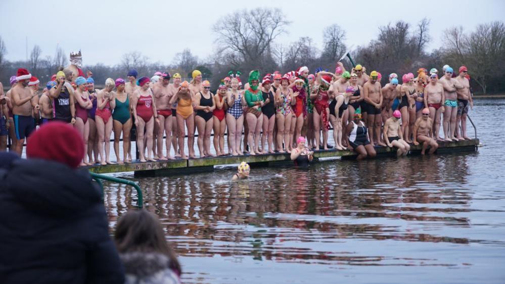 Dozens of people in swimming costumes - some in fancy dress - stand on a pontoon ready to enter a lake