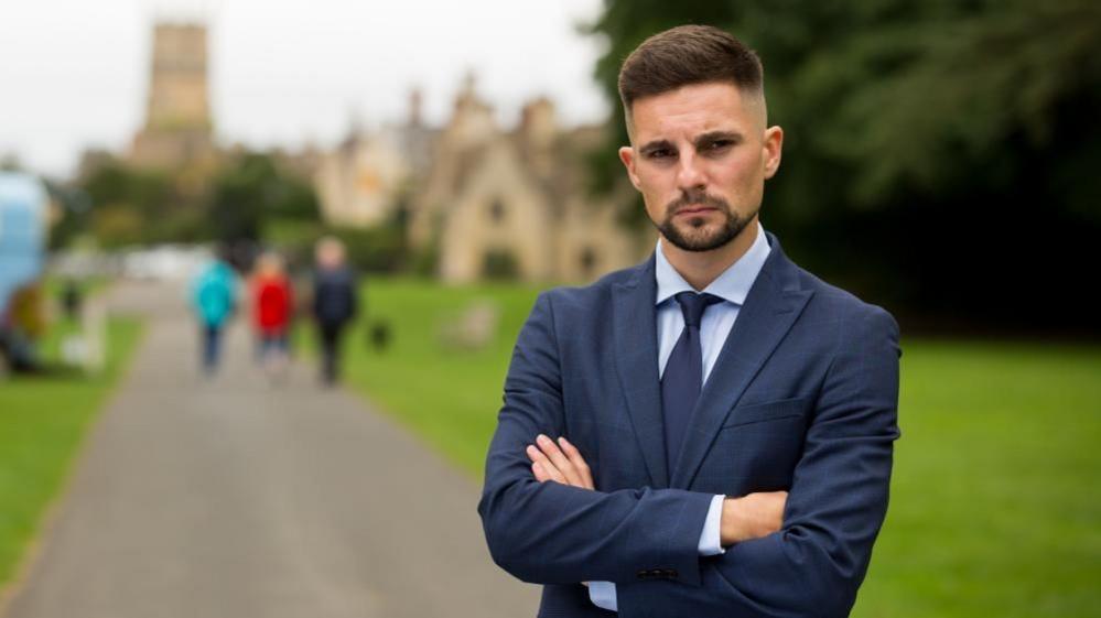 Gloucestershire County Councillor Joe Harris poses for a professional photo in Cirencester, Gloucestershire. He is wearing a blue suit and tie. He has a beard and moustache. The background is blurred with people shown walking in a park.