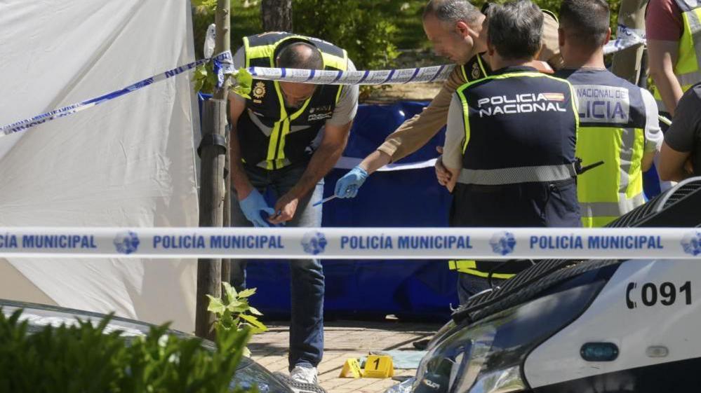 Police officers are seen looking at forensic number markers on the floor and a white tent as they search for evidence at the scene of a shooting outside the American School of Madrid (21 May 2025)