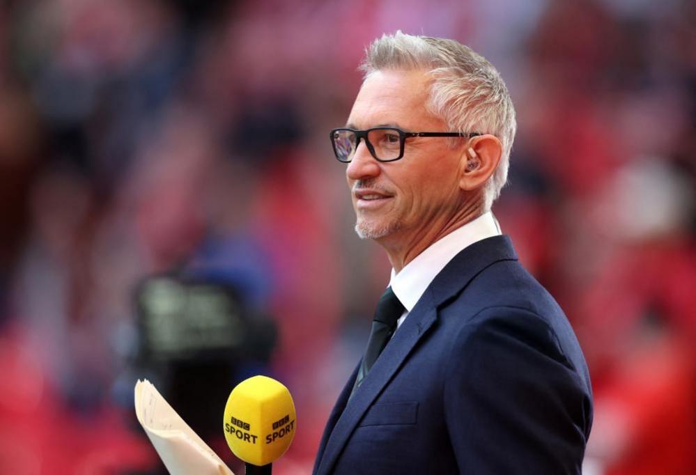 Match of the Day host Gary Lineker seen standing by the side of a football pitch before a game. He is seen side on holding a BBC Sport-branded microphone. People are visible in the stands in the background but are blurry. 