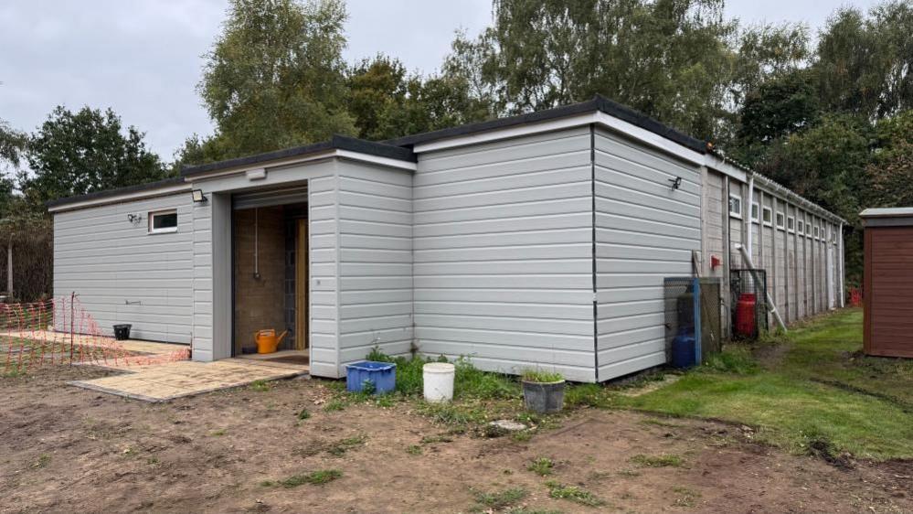 Belton Scout Hut is effectively two buildings, adjoined. A newly-clad extension with flat roof is nearest the lens, with a protruding doorway. The original part is concrete panels, with upper-light windows. Earth and grass are visible in the foreground, with trees behind.