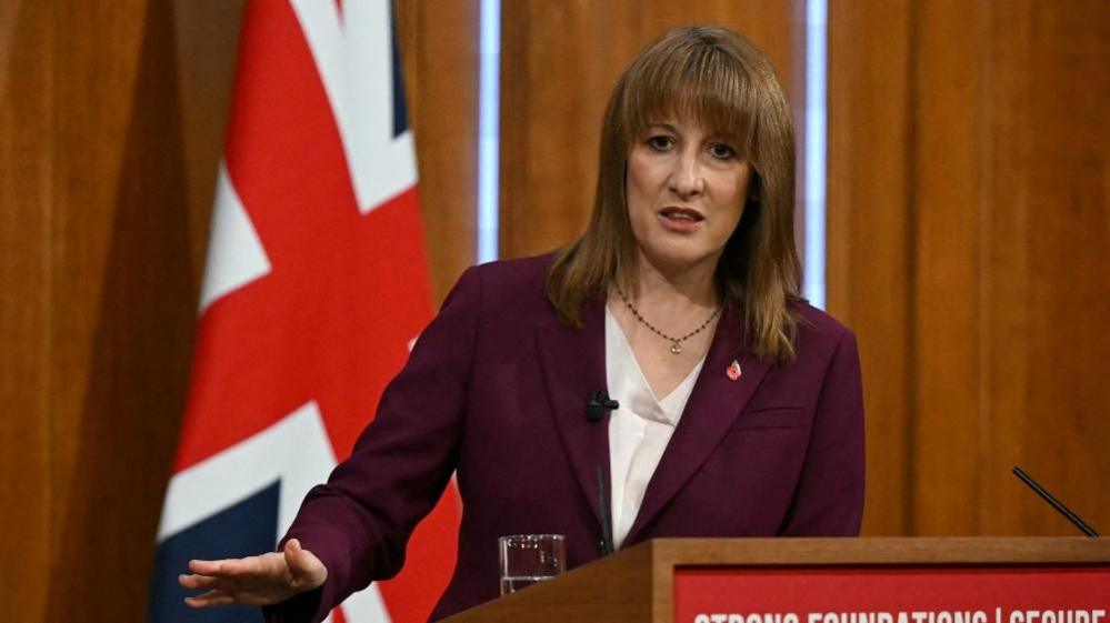 Rachel Reeves in a purple jacket looking to towards the camera as she answers a question. She is at a lectern and behind her is British flag and wood panelling. 