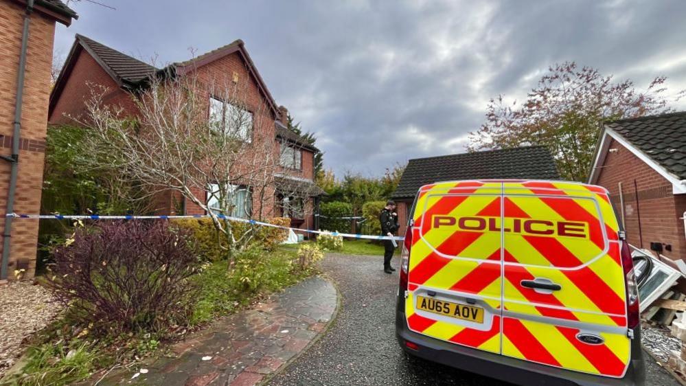 A police van is parked outside the house under investigation in Bowthorpe. The van has red and yellow high visibility chevrons and the word "police" on the back doors. The house is built of red brick and features three gables, and is bordered by other houses and garages. There is some detritus next to the van on the right side of the image, and blue and white police tape bisects the properties, creating a police cordon.