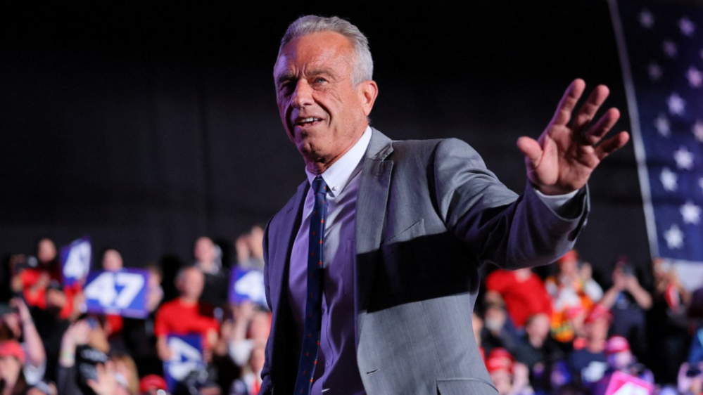 RFK Jr, who has grey hair, wears a grey suit, with a white shirt and navy patterned tie, as he waves at crowds at a Trump rally in Michigan