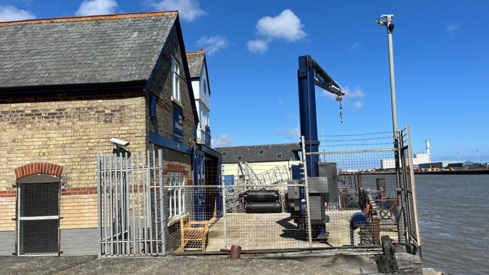 The Gorleston Lifeboat Station building is a Suffolk White brick building with red relief in bands and lintels. Fencing surrounds the building, and a blue launch Derrick crane stands on a platform outside which is used to lower the inshore Atlantic 85 lifeboat into service. Beyond the river, is the Great Yarmouth power station, and other port buildings are visible in the area.