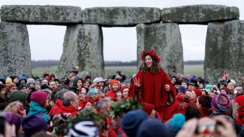 Crowds of people inside Stonehenge and a woman dressed in red sitting on someone's shoulders in the foreground