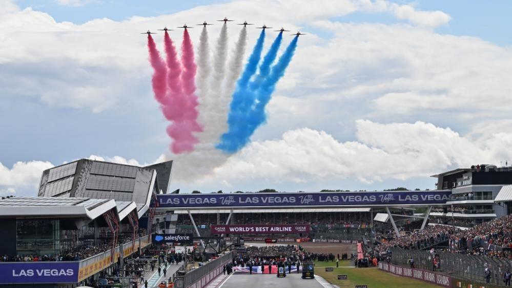 Nine red arrows with red, white and blue smoke behind them fly over stadiums at the Silverstone Circuit. The circuit is full of fans.