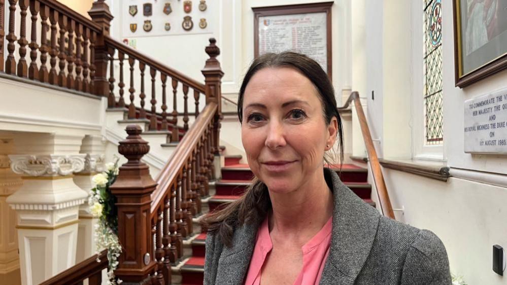Sheila Oxtoby is dressed in a pink blouse and grey jacket. She has long brunette hair tied back in a pony tail with a centre parting. She is standing on the grand staircase at Great Yarmouth Town Hall, with ornate pillars and balustrading and pictures and marble tablets mounted on the walls.