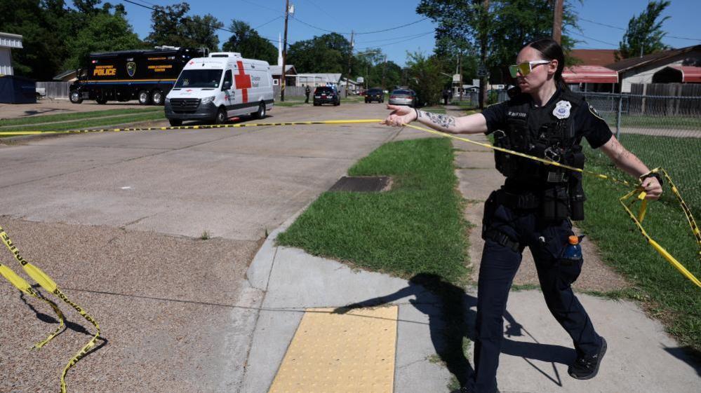 A police officer holds a police tape in a street with a police van in the distance with other vehicles, at the scene after children were killed in a mass shooting incident in Shreveport, Louisiana
