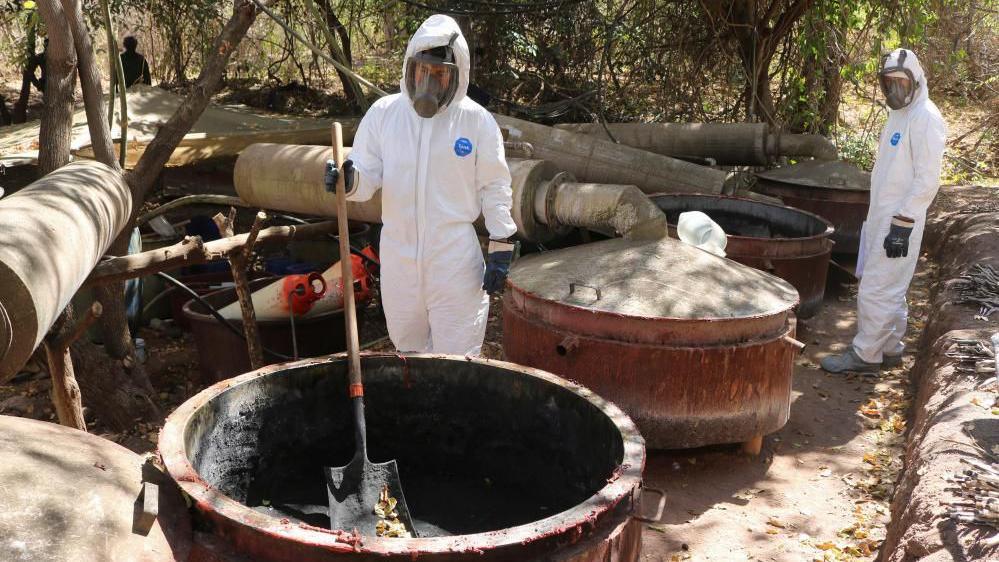 Two Mexican army personnel wearing white protective suits, full face visors and heavy-duty black gloves stand in front of large vats at a site which was used as a meth laboratory in the community of Higueras de Abuya. One of the duo is holding a spade.