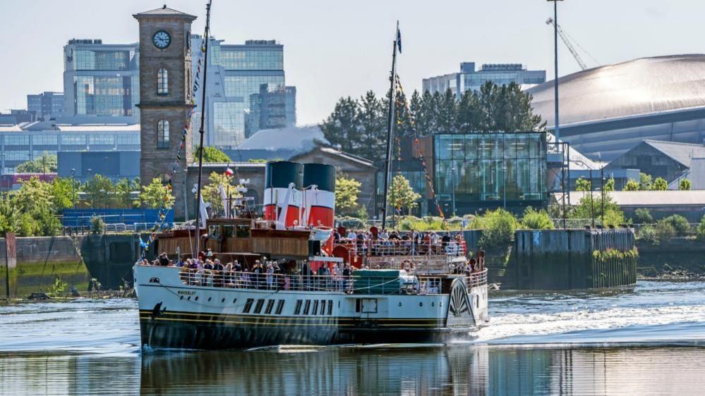 In bright sunlight, Waverley paddle steamer cruises along the River Clyde in Glasgow. Its decks are packed with people and it has two large slanted funnels in the middle, in between two masts. A clock tower can be seen beside the water in the background, as can several large, modern buildings.