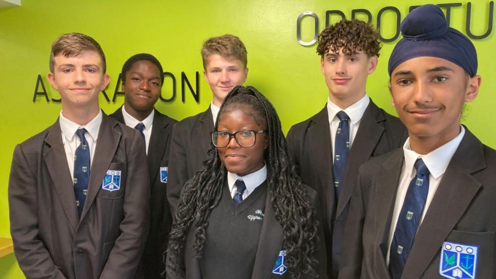 Six pupils at Wootton Upper School wearing school uniform standing in front of a green wall. All six are smiling at the camera and they are all wearing blazers and blue ties