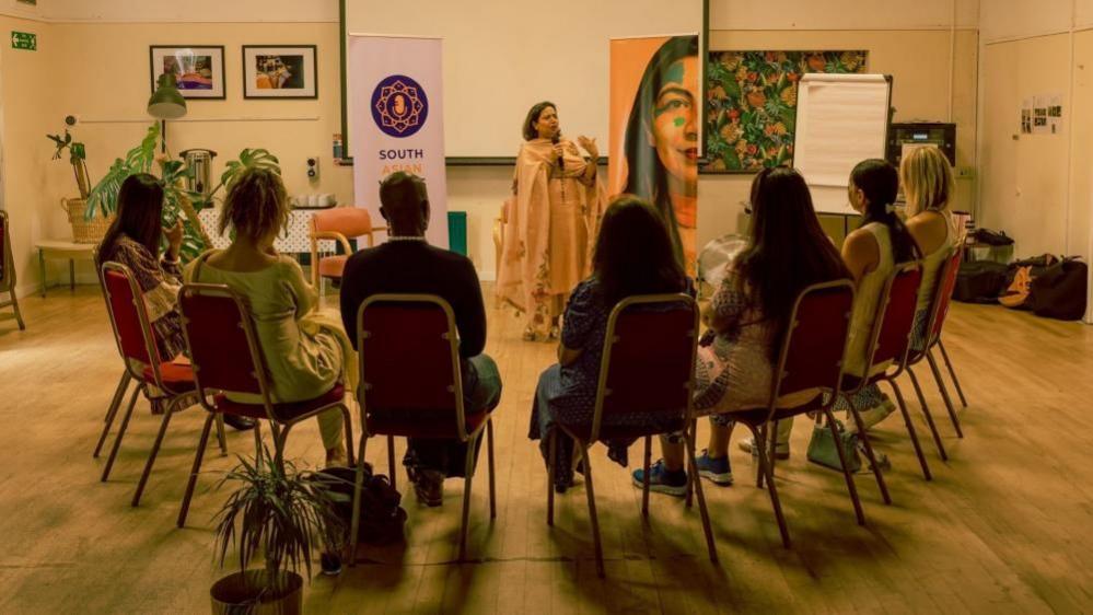 South Asian Voices Bristol CIC hosting a group discussion in a function room in Bristol. One member is speaking using a microphone. Seven other people are listening while seated in a semi circle. The woman speaking is wearing a sari.