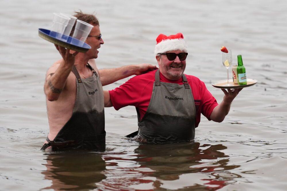 Two men in aprons stand waist-deep in the water. They both wear aprons and carry trays with drinks. One also wears a red t-shirt and a Santa hat.