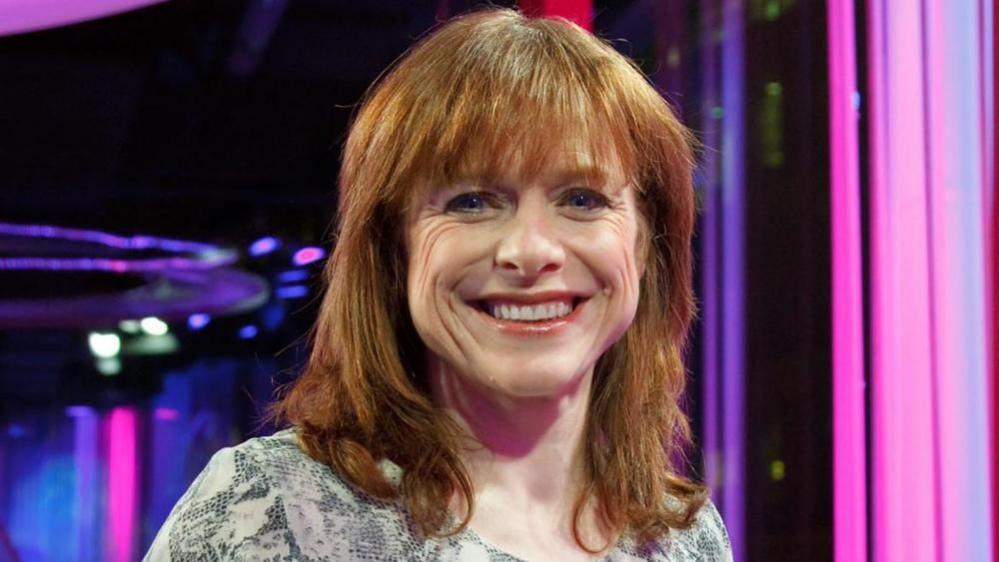 A woman with shoulder length ginger hair and a fringe smiles.