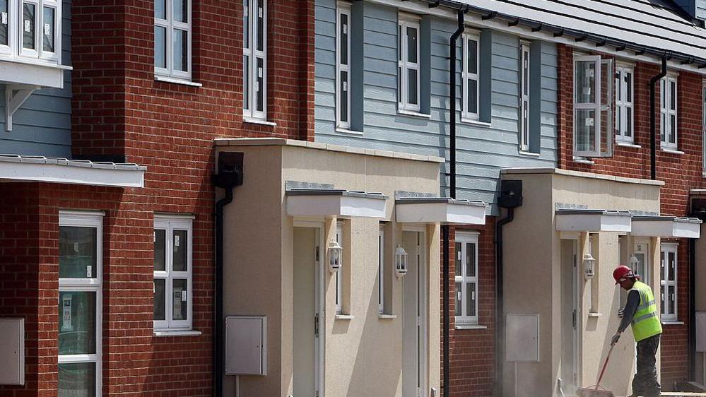 New terraced homes in Slough being prepared, with a builder sweeping outside one of the homes wearing a red hard hat and fluorescent jacket in the far right of the picture