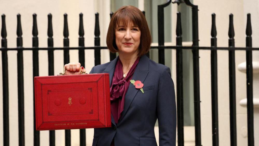 Rachel Reeves poses with the red budget box outside her office on Downing Street in London. She is wearing a navy suit with a purple blouse underneath, and has a poppy fastened to her lapel. 