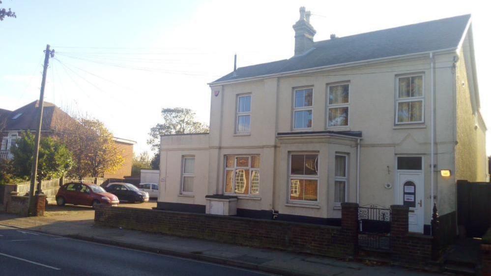 A two-storey cream-coloured house with a pitched roof and a chimney.  There is a small front garden enclosed by a low brick wall with a metal gate. To the left of the house, there is a driveway with parked cars and some trees in the background. There is a pavement and road in front of the house. 