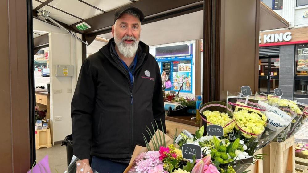 Elliot Jones with white hair and beard wearing a black gilet and black sweater.  He is standing in a market stall with orange and red roses to his right and bouquet wrapping with red ribbon to his left.