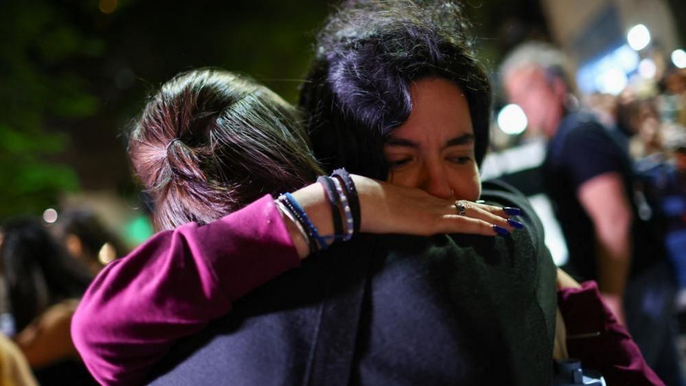 Two fans hugging outside the hotel. Facing the camera is a woman with a purple jumper on, lots of bracelets and long, dark blue nails. She has an anguished expression.