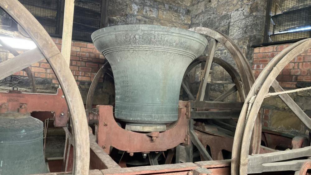 A large dark metal bell facing upwards sitting on a cast iron and wooden frame in a church tower.