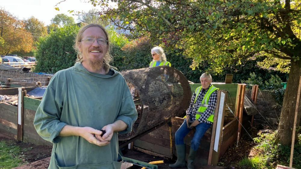 Compost mentor, Ben Bryant, who has auburn, tied back hair and matching beard, wearing a khaki fisherman's cotton tunic with pockets, standing in front of a compost tumbling machine with two volunteers sitting in the background.