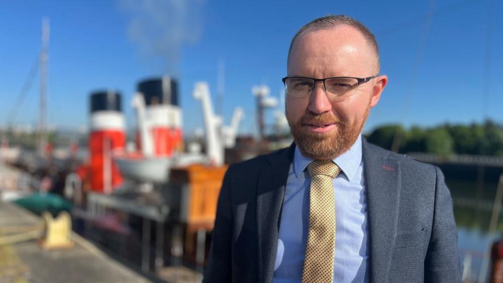 Wearing a navy jacket, blue shirt and yellow tie, Paul Semple stands in bright sunlight, with Waverley in the background. He also wears black rimmed glasses, with a neatly trimmed ginger beard. 