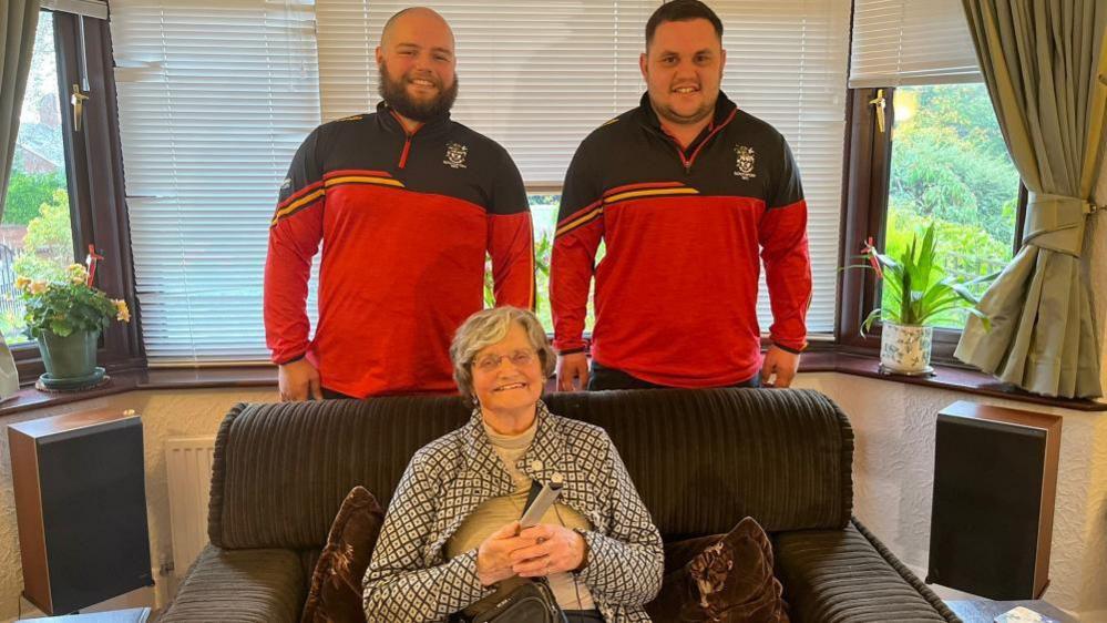 Two men in rugby tops stand behind an old woman who is sitting in a large armchair in front of a large bay window. She is holding a TV remote, has grey hair and glasses and a patterned jacket on.