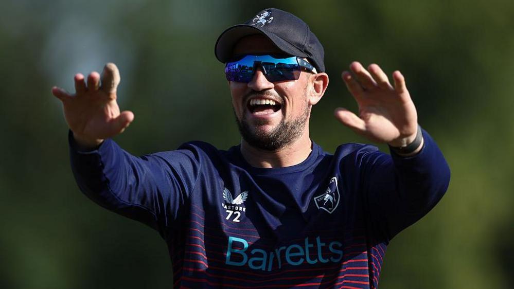 Harry Finch smiles with both arms raised in front of him, wearing a blue baseball cap, dark sunglasses and a blue cricket kit