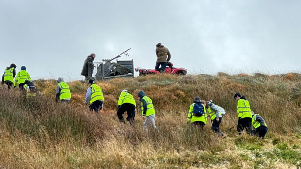 A line of teenagers in high viz jackets walking up a hill, in the backround are two men, one on a red quead towing the wreck. 