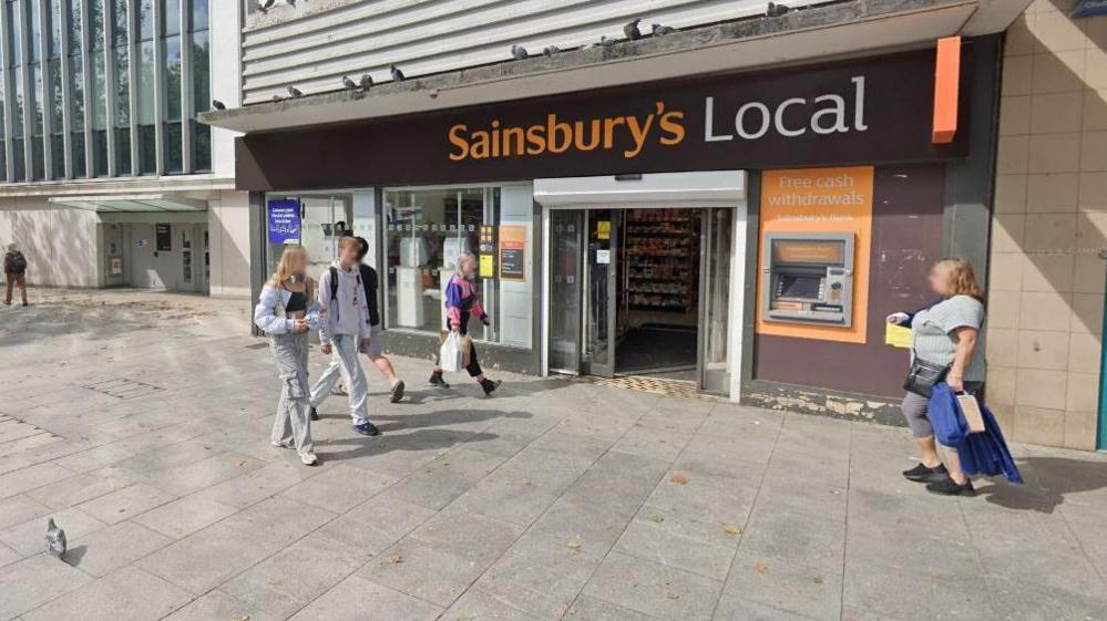 Google Street View image of people walking on the pavement outside a Sainsbury's Local shop on Sidwell Street in Exeter. Five people are walking past it. One of the people - a woman wearing a pink and navy blue rugby top looks like she is about to enter the shop. A cash point is on the exterior wall of the store, which has large windows on the front and double electric doors.