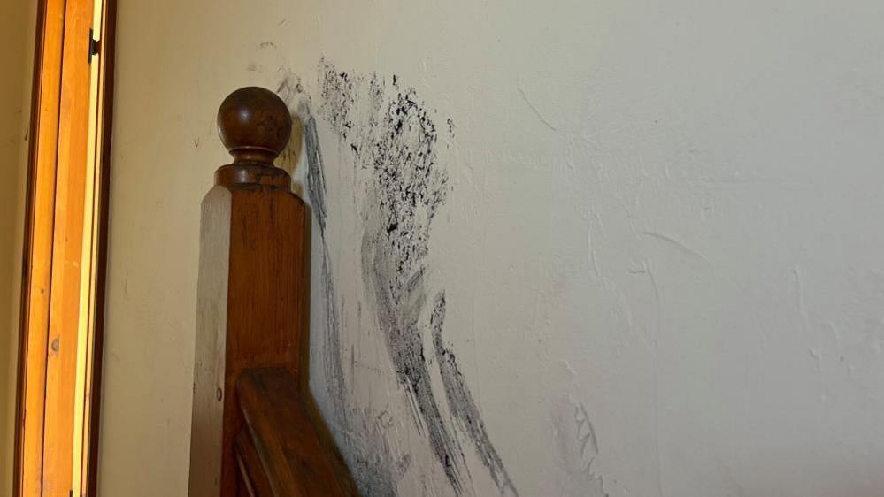 Soot stain marks next to the top floor newell post at the top of a staircase. The wall is white rough plastered, and the newell post is made of wood and features a ball on the top.