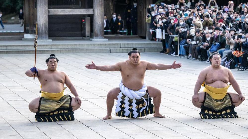 Hoshoryu, topless and in ceremonial dress from the waist down, crouched with two other wrestlers either side of him