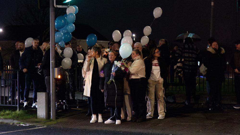 A group of people standing at the side of a road at a pedestrian crossing. They are holding blue and white balloons and some are holding flowers. 