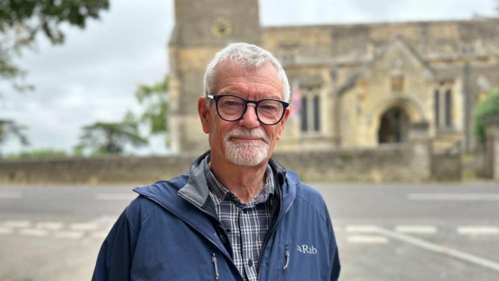 Trevor Higgs with short white hair and beard wearing a blue jacket and standing at a road junction with a church behind him