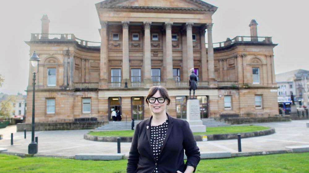 A woman, wearing a black blazer and black and white polka dot dress, stands in front of an impressive neoclassical building with tall columns and a triangular pediment, set in a landscaped square with a statue and green lawn.