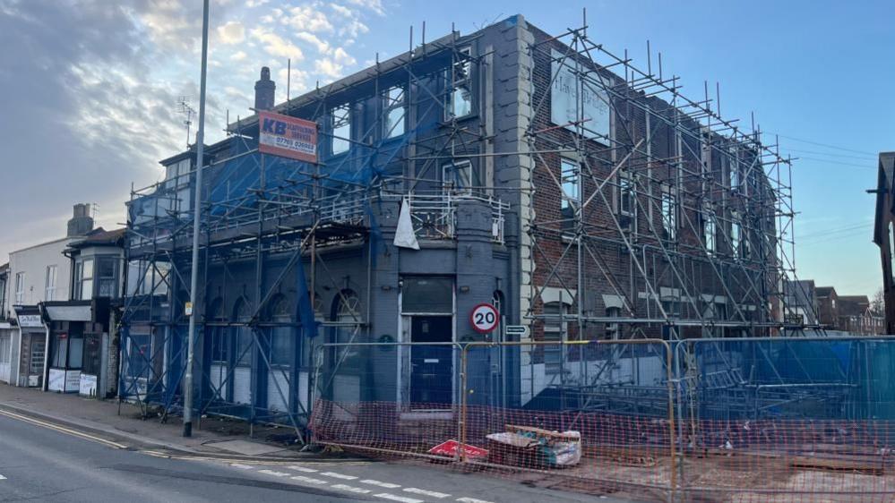 The former Haven Bridge pub, shrouded in scaffolding and blue netting to keep dust and debris within. A terrace of shops is in the far left of the image, and the blocked entrance to Steam Mill Lane on the right.