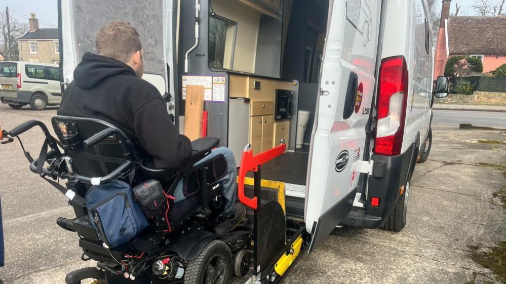 A man is sat in a powered wheelchair. He is facing away from the camera, has blonde hair and is wearing a black hoodie. The chair is on a lift attached to the back of a white van. The lift has bright red handlebars and the edges are bright yellow. Around the van, the floor is concrete. The sky can just be seen at the sides, and it is grey. 