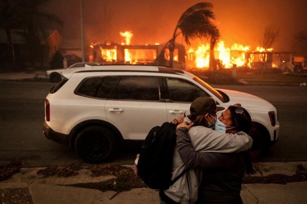Two women are visibly upset as they embrace, with fires raging in the background. They are wearing face masks and dark clothes and one of them carries a rucsac. A white car stands behind them. The fires have already largely destroyed the buildings on the other side of the road and damaged trees sway in the wind.